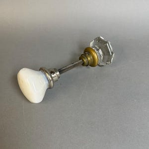 May include: A door knob set featuring one smooth, opaque white knob and one faceted, clear glass knob. The two knobs are connected by a central metal spindle, with a brass-toned collar at the base of the glass knob and a silver-toned collar at the base of the white knob.