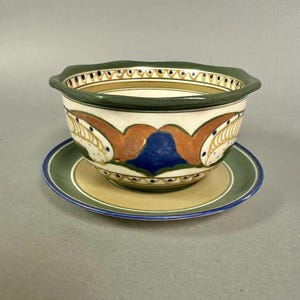 May include: A ceramic bowl with a matching saucer. The bowl has a scalloped rim and is painted with a cream, green, blue, and orange design. The saucer is green with a blue rim. The bowl sits on the saucer.
