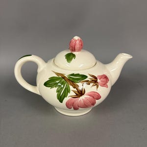 May include: Cream-colored ceramic teapot with a floral design. The teapot features pink flowers, green leaves, and brown stems. The lid has a pink bud-shaped handle. The handle and spout are curved.
