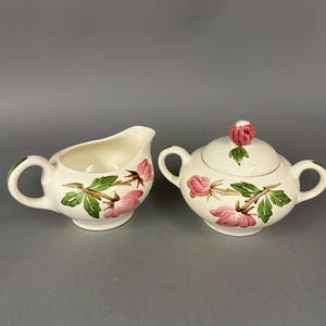 May include: Cream-colored ceramic creamer and sugar bowl set. The set features pink rose and green leaf designs. The sugar bowl has a lid with a pink rose knob. The creamer and sugar bowl have curved handles.