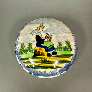 May include: Round ceramic trinket box with a hand-painted scene. The lid features a seated figure in traditional attire holding flowers, set against a landscape with trees and a cloudy sky. The box has a blue and white floral border.