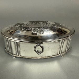 May include: An oval silver trinket box with an ornate floral design on the lid and a beaded edge. The box has a hinged lid with a decorative handle.