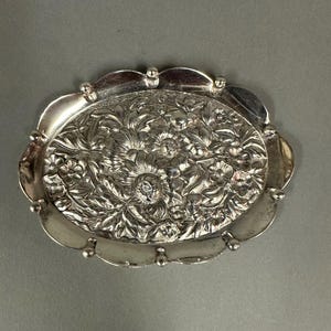 May include: A silver oval tray with a raised floral design. The tray has a scalloped edge and is decorated with small beads around the rim.