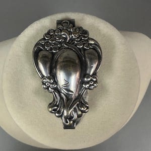 May include: A silver-toned, Art Nouveau-style brooch with floral and flowing leaf designs. The brooch features a central, elongated shape with three rounded lobes and small flower clusters. The metal has a slightly aged appearance.