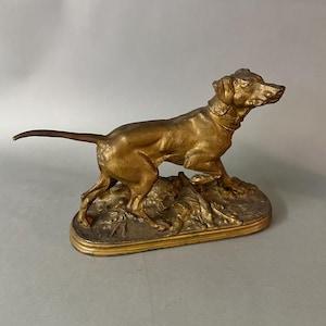 May include: A golden-colored dog sculpture in a hunting pose. The dog is on a rectangular base with detailed foliage. The sculpture has a smooth, metallic finish and is likely made of bronze or a similar material.