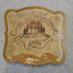May include: A ceramic plate with a vintage design featuring a house, trees, and the text "Chateau Boncoeur Chardonnay 1896 Burgundy".