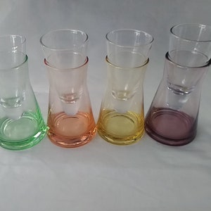 May include: Four clear glass shot glasses with colored bases. The glasses are stacked on top of each other, with the top glass being clear and the bottom glass being colored. The colors of the bases are green, orange, yellow, and purple.
