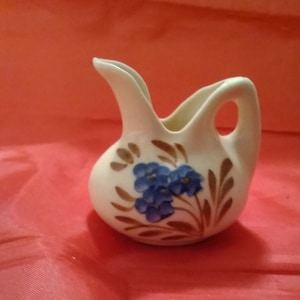May include: A small white ceramic pitcher with a blue floral design. The pitcher has a handle and a spout.