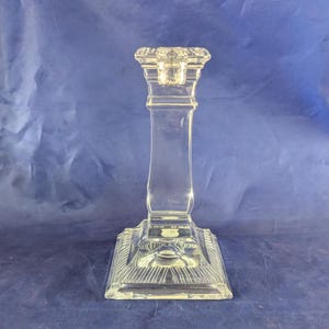 May include: Clear glass candlestick holder with a square base and a detailed, ornate design. The candlestick has a tall, slender stem and a top designed to hold a single candle. The glass is transparent, allowing light to pass through.