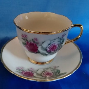 May include: A white teacup and saucer set with a gold rim and floral design featuring pink and white roses.