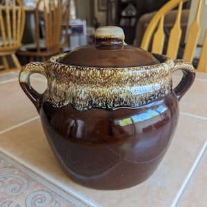 May include: A brown glazed ceramic crock with a lid. The crock has a white rim and a speckled glaze.