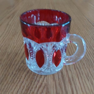 May include: A clear glass cup with a red rim and red accents. The cup has a clear glass handle.