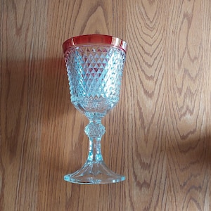 May include: A clear glass goblet with a red rim. The goblet has a diamond pattern on the bowl and a decorative stem and base. The glass is sitting on a wooden surface.