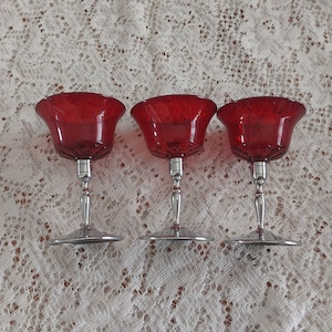 May include: Three red glass stemmed cocktail glasses with silver bases. The glasses are arranged in a row on a white lace tablecloth.