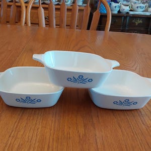 May include: Three white, square casserole dishes with blue floral designs. Each dish has a handle on either side. The dishes are stacked on a wooden table.