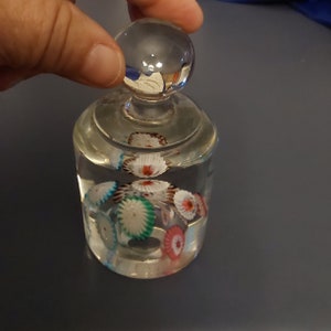 May include: A clear glass bottle stopper with a round top and a clear glass base. The base contains colorful glass flowers.