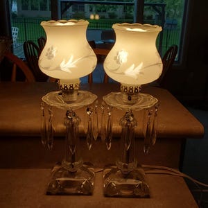 May include: Two vintage table lamps with frosted glass shades featuring floral designs. The lamps have crystal bases and dangling crystal accents. The shades are illuminated, casting a warm glow. The lamps are placed on a brown surface.