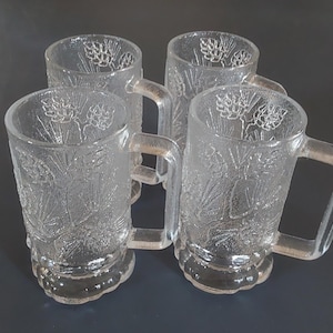 May include: Four clear glass beer mugs with handles. Each mug has a textured wheat stalk design. The mugs are arranged in a square formation, showcasing their transparent nature and decorative details.