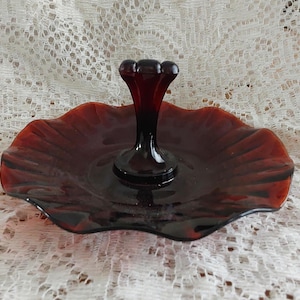 May include: A vintage ruby red glass serving dish with a central stem. The dish has a ruffled edge and a dark, glossy finish. The stem is topped with three rounded accents.