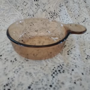 May include: A clear, amber-colored glass bowl with a handle. The bowl has a rounded shape and a dark rim. The handle is small and curved, extending from the side of the bowl. The bowl is likely for cooking or serving.
