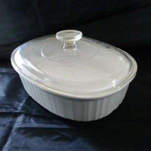 May include: Oval, white casserole dish with a clear glass lid and a small, round handle. The dish has vertical ribbed detailing on the sides. A classic piece for baking and serving.