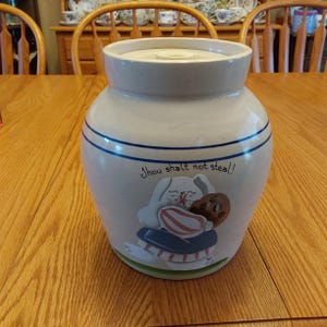 May include: A white ceramic cookie jar with a blue and white striped design. The jar has a lid and features a painted image of a bunny holding a cookie. The text "Thou shalt not steal!" is written on the jar.