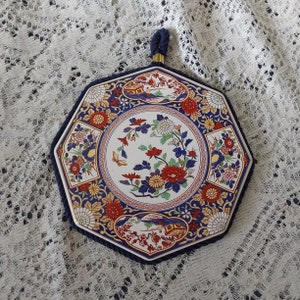 May include: Octagonal ceramic trivet with a blue and white floral design. The trivet has a gold trim and a blue tassel.