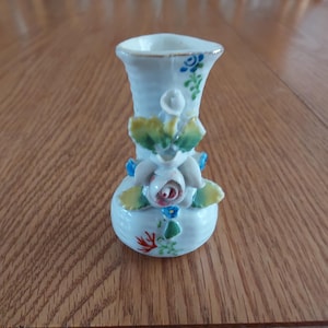 May include: A small white porcelain vase with a floral design. The vase has a raised, three-dimensional flower design with white, pink, blue, and green accents.