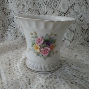 May include: A white ceramic vase with a scalloped rim and a floral design. The vase features a bouquet of pink, yellow, and purple flowers with green leaves.