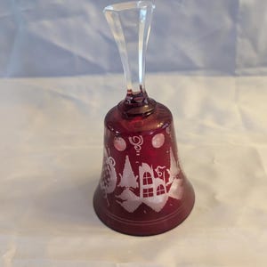 May include: A ruby red glass bell with a clear handle. The bell features a white etched design of a village scene with houses, trees, and decorative swirls. The bell is approximately 5 inches tall.