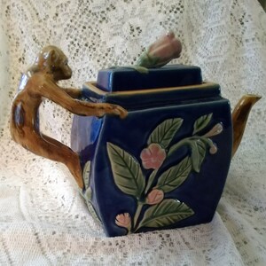 May include: A blue ceramic teapot with a monkey handle and floral design. The lid has a pink flower on top.
