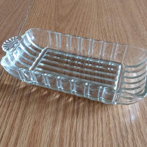 May include: A rectangular, clear glass dish with a ribbed design and decorative handles. The dish is likely for serving or display, and sits on a wooden surface.