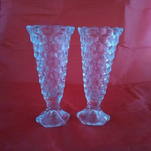 May include: Two clear glass bud vases with a textured diamond pattern. The vases are on a red background.