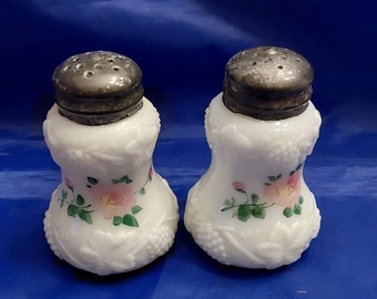 May include: A pair of white, vintage salt and pepper shakers with silver-colored lids. The shakers feature a floral design with pink roses and green leaves. The base of each shaker has a grape cluster pattern.