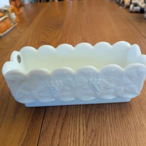 May include: White milk glass dish with a scalloped edge and a grapevine design. The dish is perfect for serving snacks or holding small items.