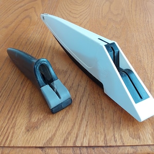 Op de afbeelding: Twee keukengereedschappen worden weergegeven op een houten oppervlak. E&eacute;n is een witte en zwarte messenslijper en de andere is een zwarte en grijze handgreep. De gereedschappen zijn ontworpen voor het slijpen van messen en andere snijgereedschappen.