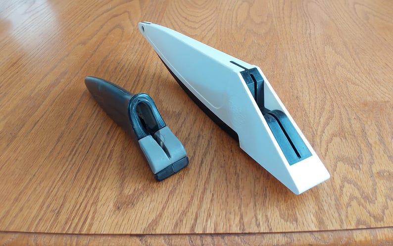 Op de afbeelding: Twee keukengereedschappen worden weergegeven op een houten oppervlak. E&eacute;n is een witte en zwarte messenslijper en de andere is een zwarte en grijze handgreep. De gereedschappen zijn ontworpen voor het slijpen van messen en andere snijgereedschappen.