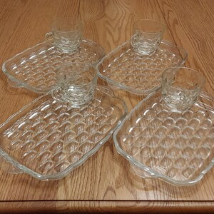 May include: Four clear glass teacups and saucers. Each saucer has a textured, bubble-like pattern. The teacups sit in the center of the saucers. The set is on a wooden surface.