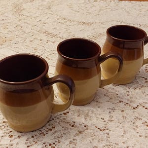 May include: Three ceramic mugs with a brown and tan color scheme. Each mug has a rounded body, a dark brown interior, and a matching handle. The mugs are arranged in a row on a white lace tablecloth.