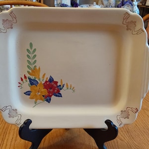 May include: Cream-colored rectangular serving platter with scalloped edges and corner handles. The platter features a floral design in yellow, red, and blue. Decorative accents are on the corners. The platter is displayed on a black stand.