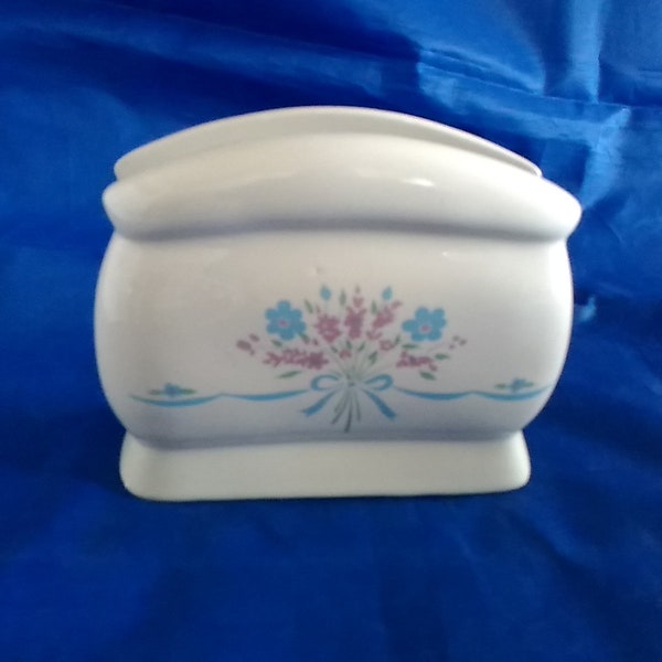 A white ceramic napkin holder with a floral and ribbon design in lilac and blue with greenery.  Dish 1414