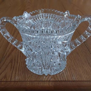 May include: Clear, cut-glass sugar bowl with two handles. The bowl features intricate geometric patterns and a scalloped rim. The handles are curved and integrated into the design. The bowl is made of clear glass and sits on a wooden surface.