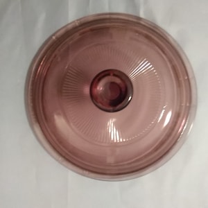 May include: A clear glass lid with a purple tint and a raised knob in the center. The lid has a concentric circle pattern.
