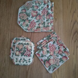 May include: Set of six fabric placemats and two pot holders with a fruit print design. The placemats are rectangular and the pot holders are square. The fabric is a cream color with a green, pink, and brown fruit print.