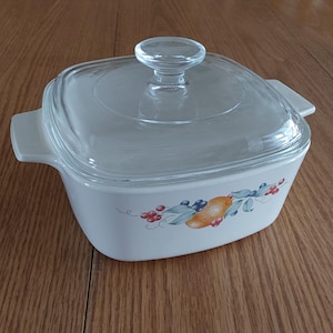 May include: Square, off-white casserole dish with a clear glass lid and handle. The dish features a fruit and berry design in shades of orange, blue, and red. The dish is sitting on a wooden surface.