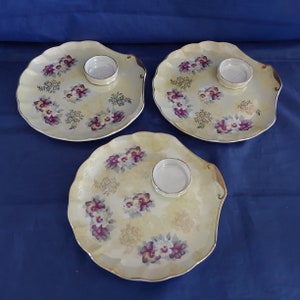 May include: Three small, white ceramic serving plates with a scalloped edge. Each plate has a small, round, white dish in the center and a floral design with purple, yellow, and gold accents.