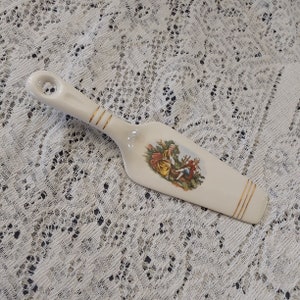 May include: A white ceramic cake server with a gold trim and a painted scene of two people in a garden.