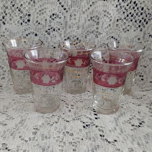 May include: A set of five clear glass shot glasses with a decorative band of pink and white floral designs. The glasses have a classic shape and are arranged on a white lace tablecloth.