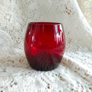 May include: A small, round, ruby red glass bowl. The bowl is sitting on a white lace tablecloth.