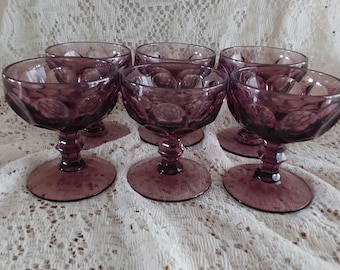 May include: Six vintage amethyst-colored stemmed glasses. Each glass features a round bowl with a textured pattern and a short, decorative stem. The glasses are arranged on a white lace tablecloth.